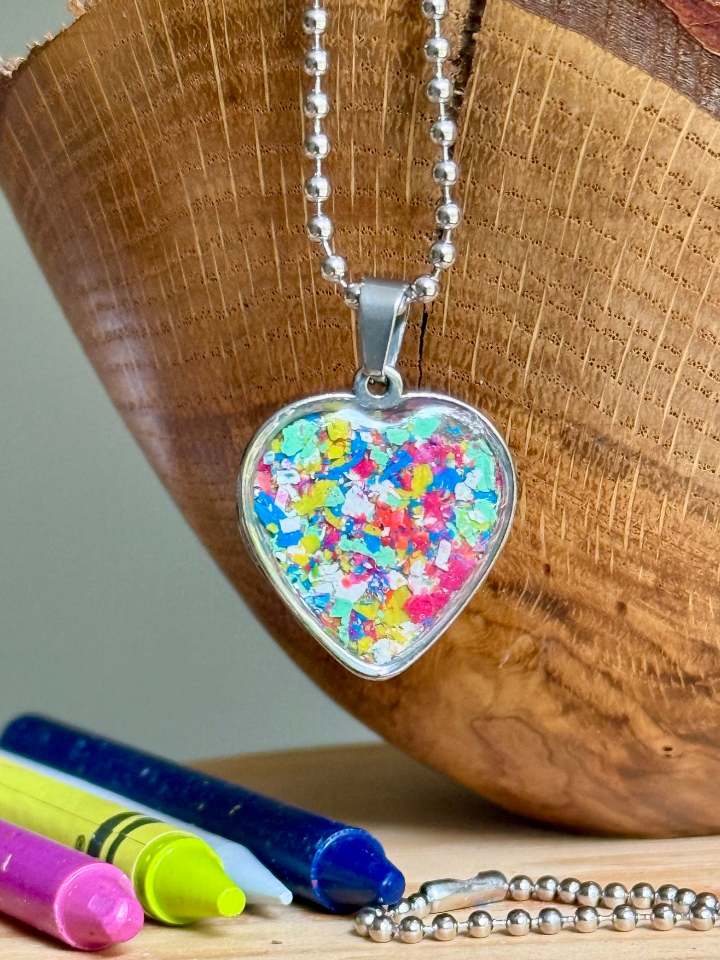 A heart-shaped pendant necklace filled with colorful confetti flakes, hanging from a silver ball chain. The pendant is displayed on a rustic wooden surface next to crayons.