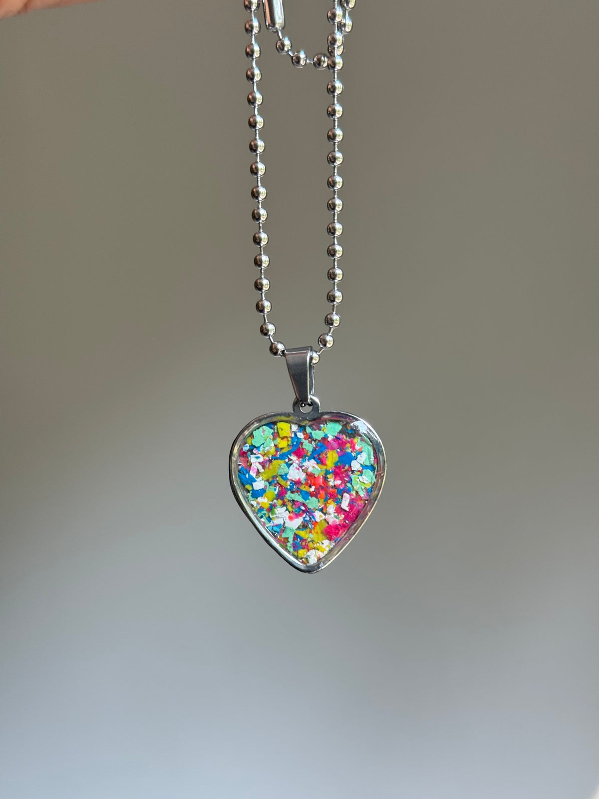 A stainless steel heart-shaped pendant necklace filled with colorful crayon, hanging from a silver ball chain against a soft gray gradient background.