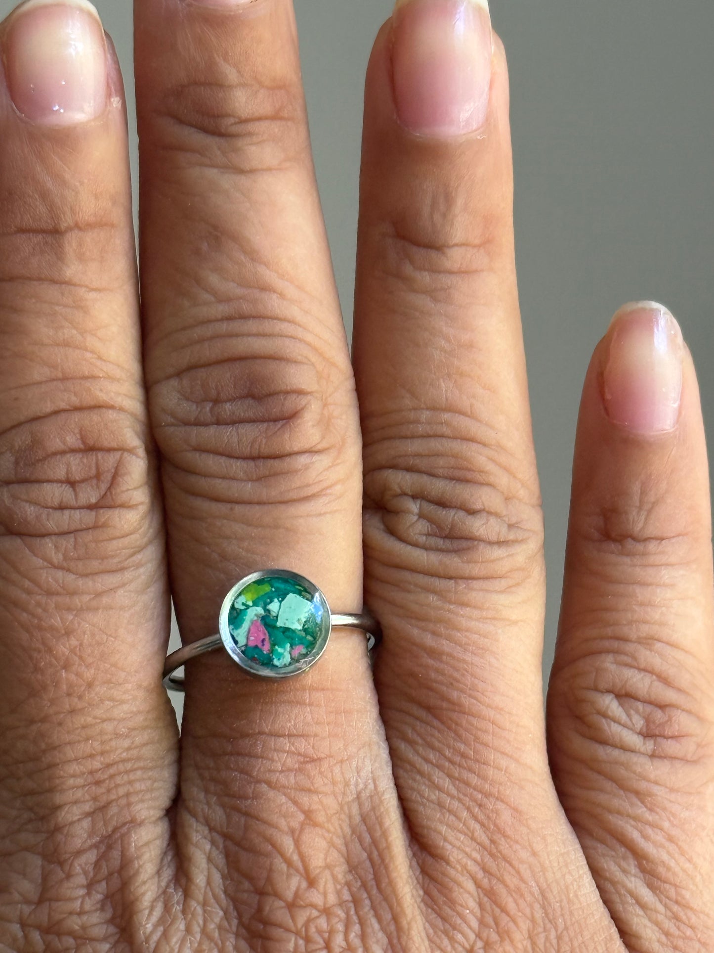 A person wearing a dainty stainless steel watermelon-toned crayon jewelry ring on their finger, showcasing a unique colorful mosaic of pink, teal, and green fragments.