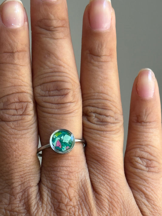 A person wearing a dainty stainless steel watermelon-toned crayon jewelry ring on their finger, showcasing a unique colorful mosaic of pink, teal, and green fragments.