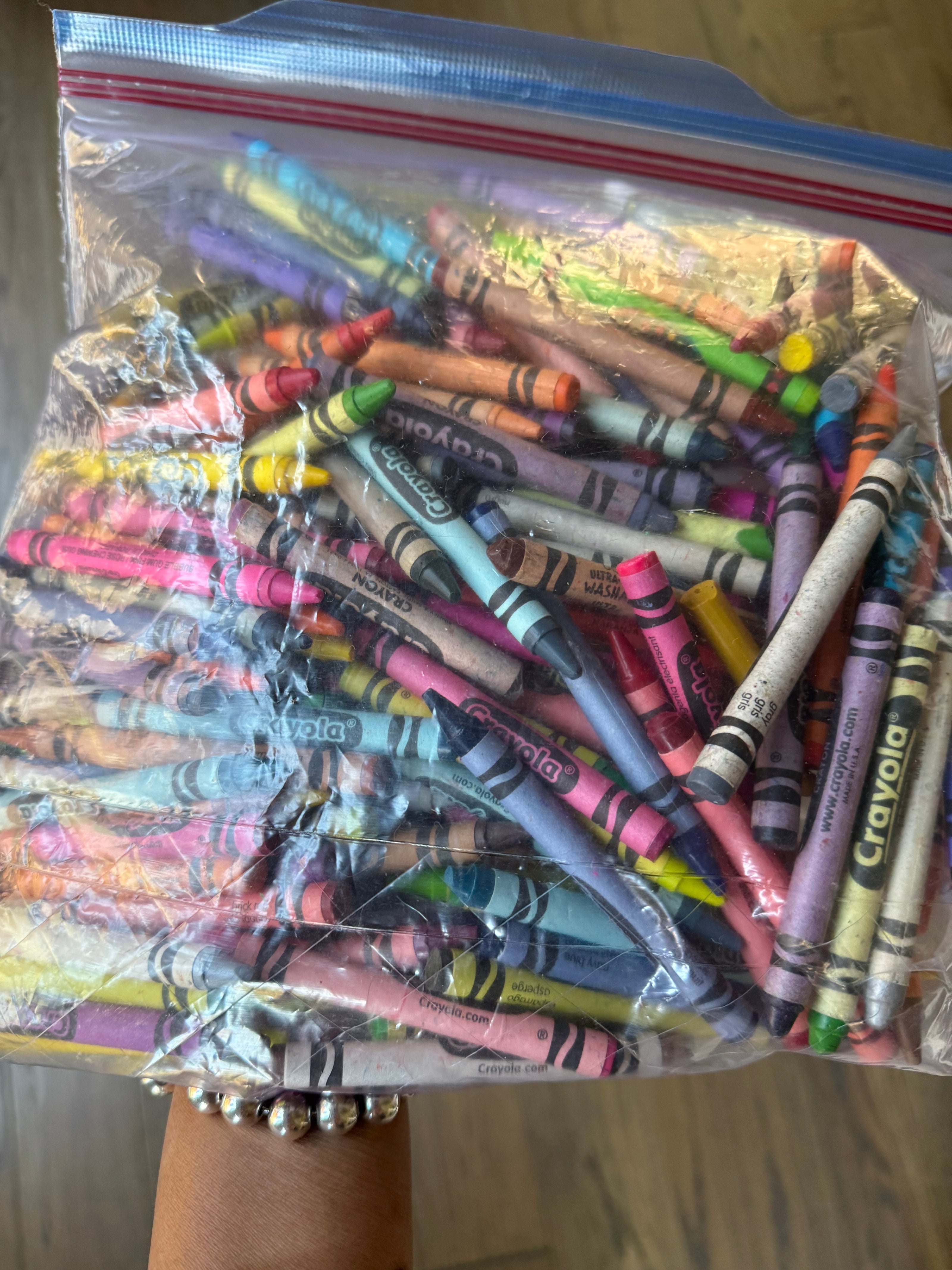 A clear plastic zip-top bag filled with many crayons in assorted colors, held up by a person's hand against a wood floor background.