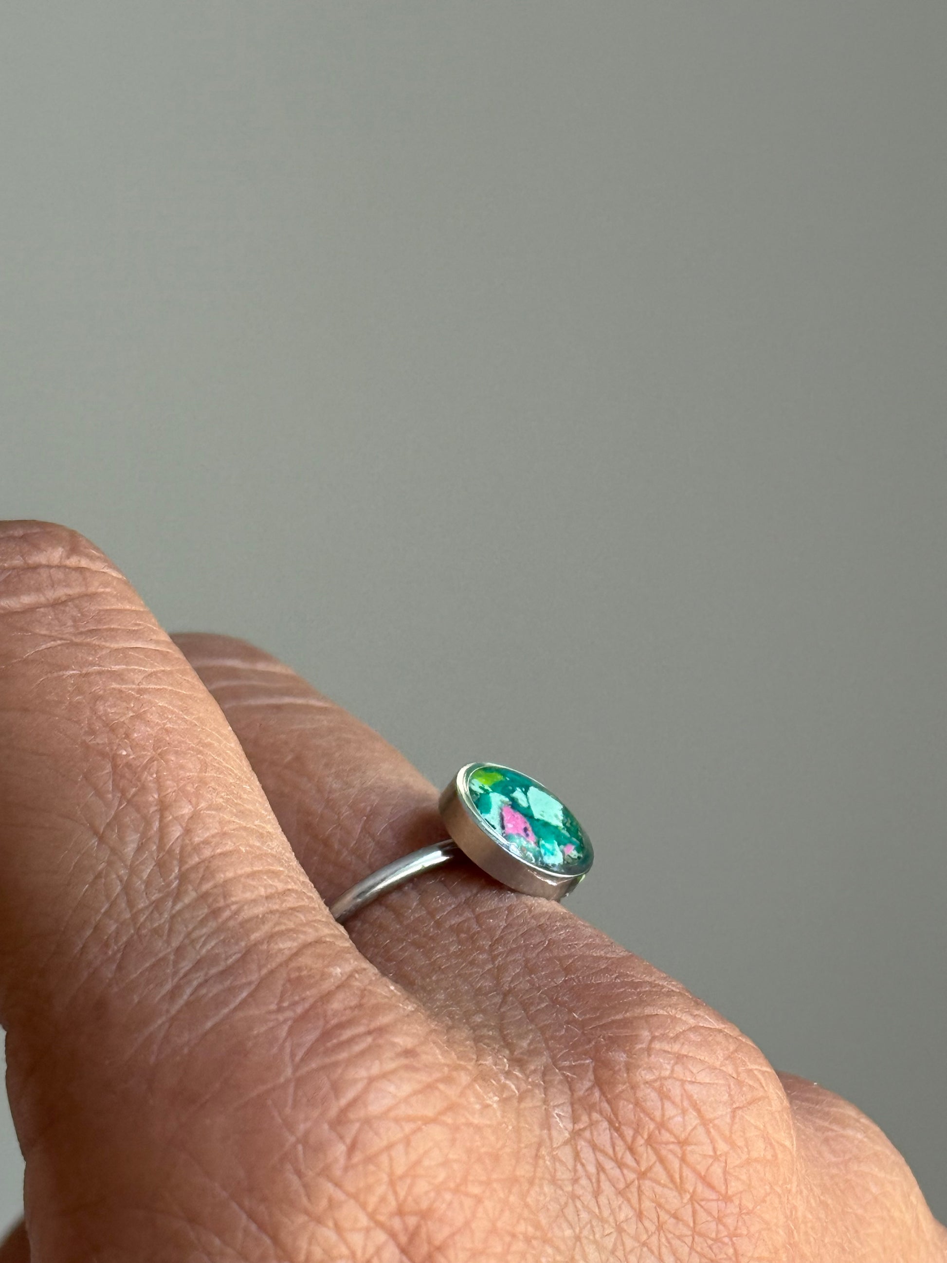 A minimalist stainless steel jewelry ring featuring a colorful, one-of-a-kind center filled with a textured mix of pink, green, and teal crayon shavings