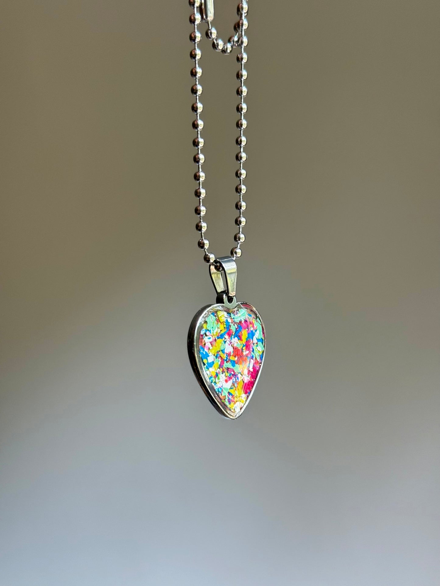 A stainless steel heart-shaped pendant necklace filled with multicolored crayon shavings.  Crayon jewelry bt Crayonfetti. By mother-daughter.