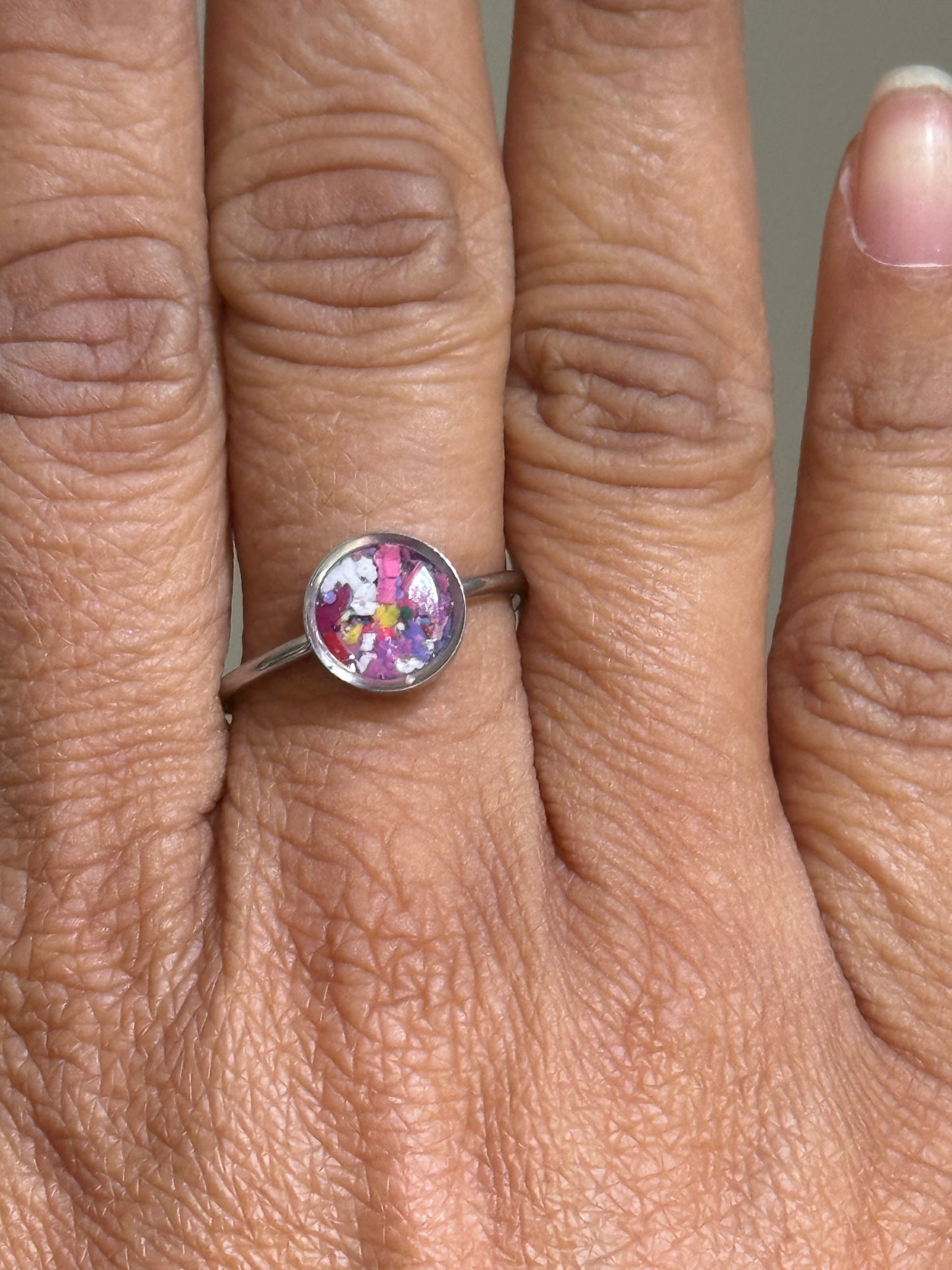 Close-up of a delicate stainless steel ring filled with a whimsical burst of colorful crayonfetti against a cloud-like purple and white background.