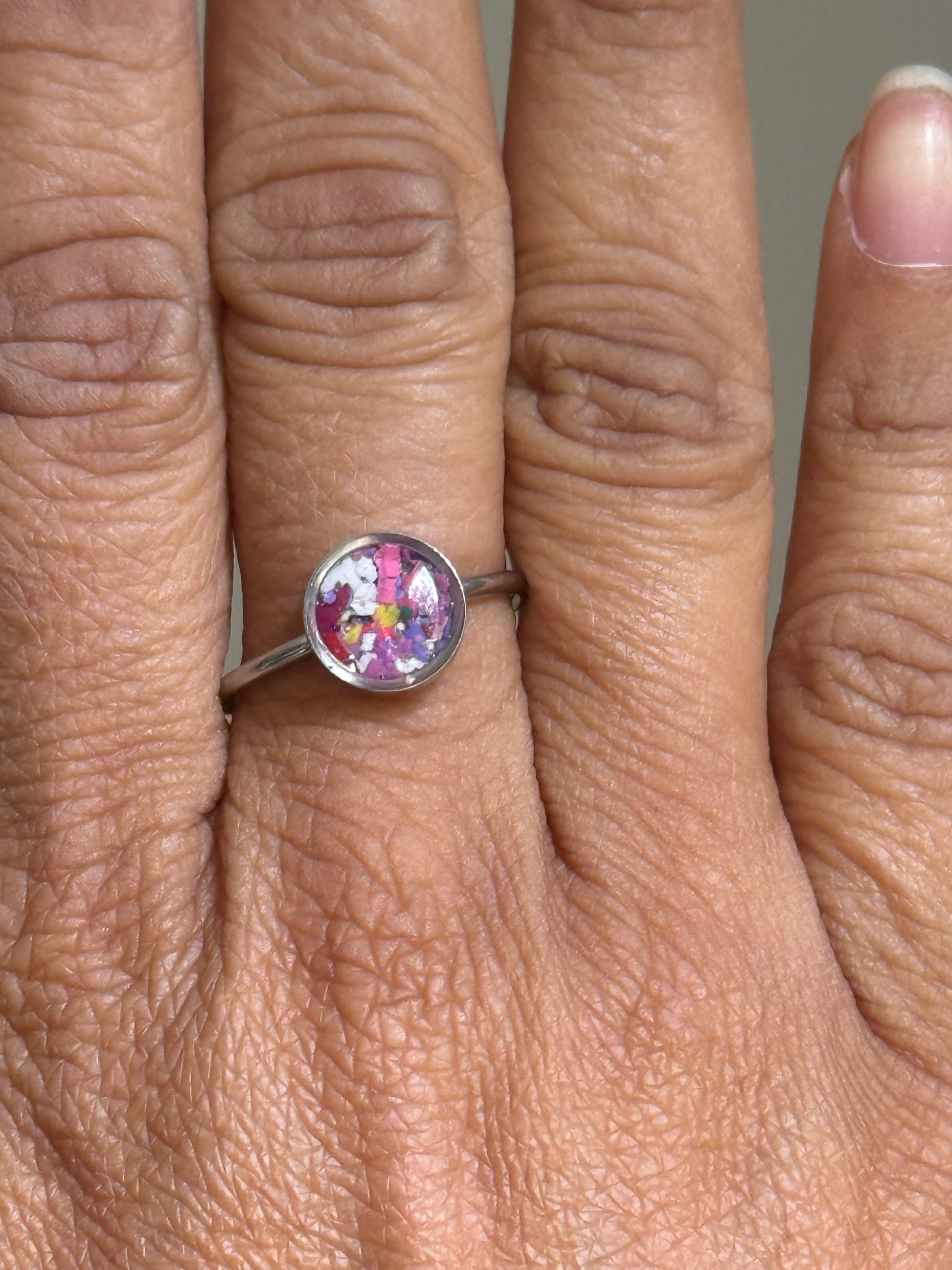 Close-up of a delicate stainless steel ring filled with a whimsical burst of colorful crayonfetti against a cloud-like purple and white background.