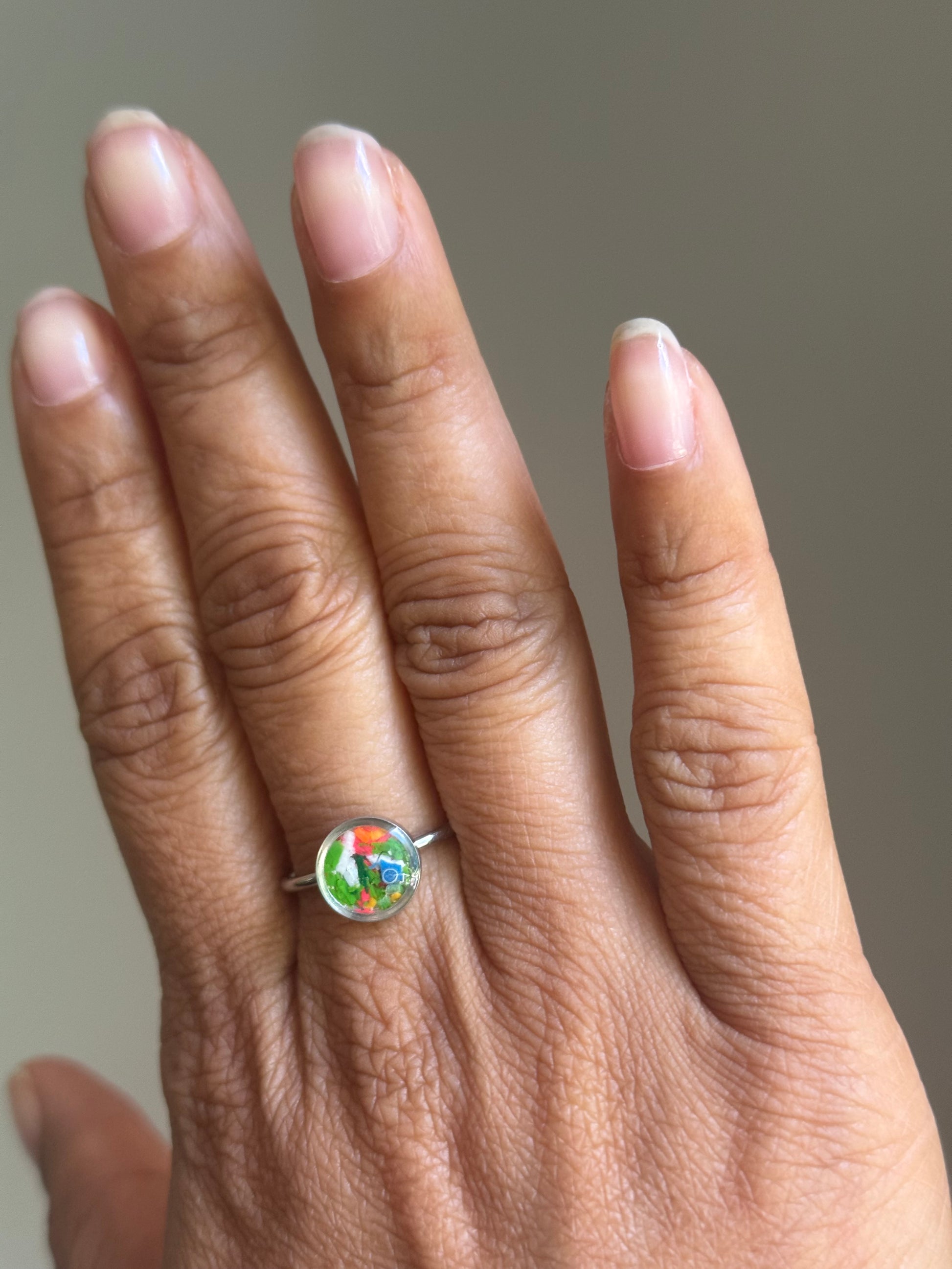 A person wearing a minimalist stainless steel Sunkist ring on their finger, styled as a dainty stacking ring alongside other colorful jewelry.