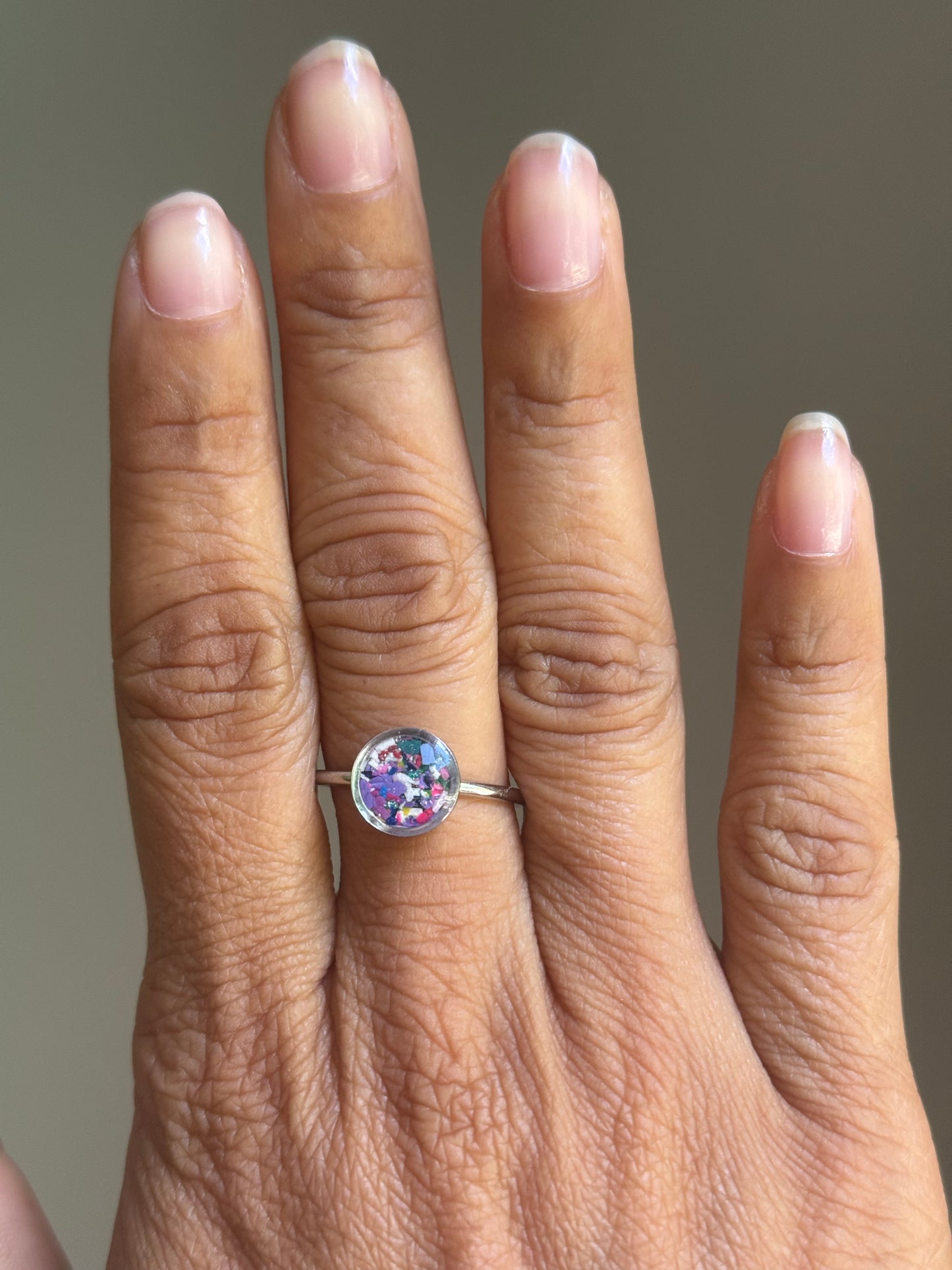 A person wearing a minimalist stainless steel Lavender Mist ring on their finger, styled as a dainty stacking ring alongside other colorful jewelry.