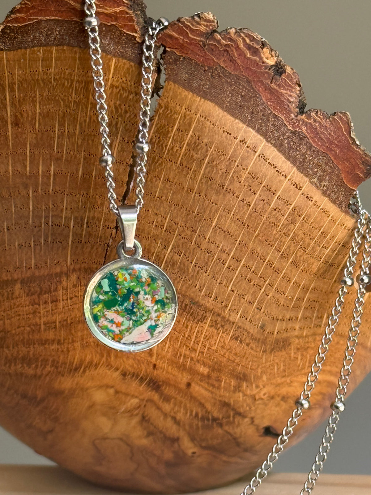 Close up of dime-sized round pendant filled with blue green orange and pink reclaimed crayon jewelry fragments in steel bezel.
