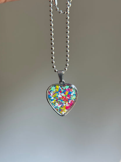 A stainless steel  heart-shaped pendant necklace filled with colorful crayon, hanging from a silver ball chain against a soft gray gradient background.