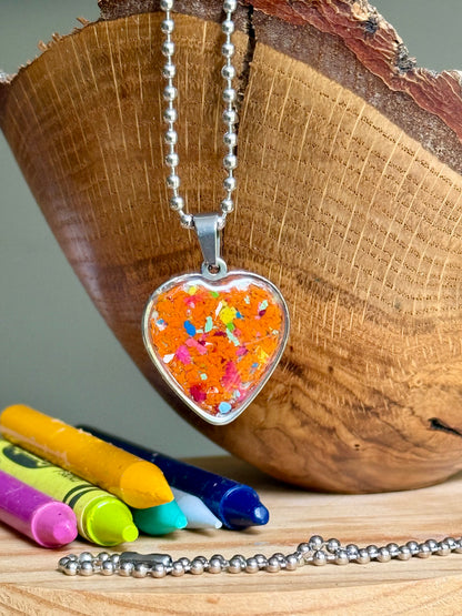 Close-up of handmade orange crayon heart jewelry showing colorful recycled crayon shavings preserved in resin. Crayon Jewelry by Crayonfetti