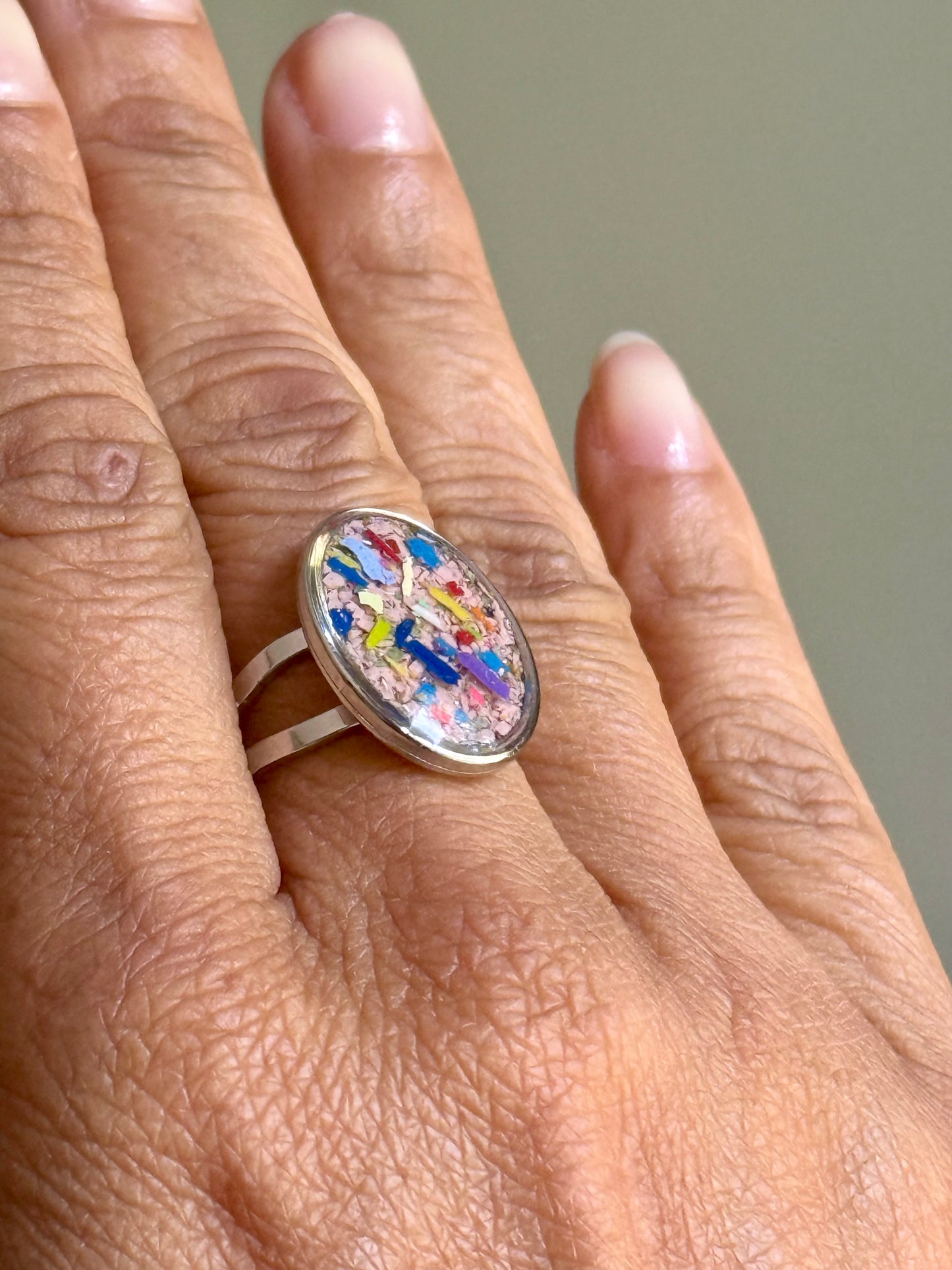 A close-up of a hand wearing a stainless steel oval ring with a colorful, abstract mosaic-like oval cabochon. Crayonfetti