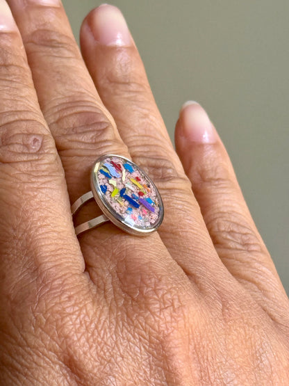A close-up of a hand wearing a stainless steel oval ring with a colorful, abstract mosaic-like oval cabochon. Crayonfetti