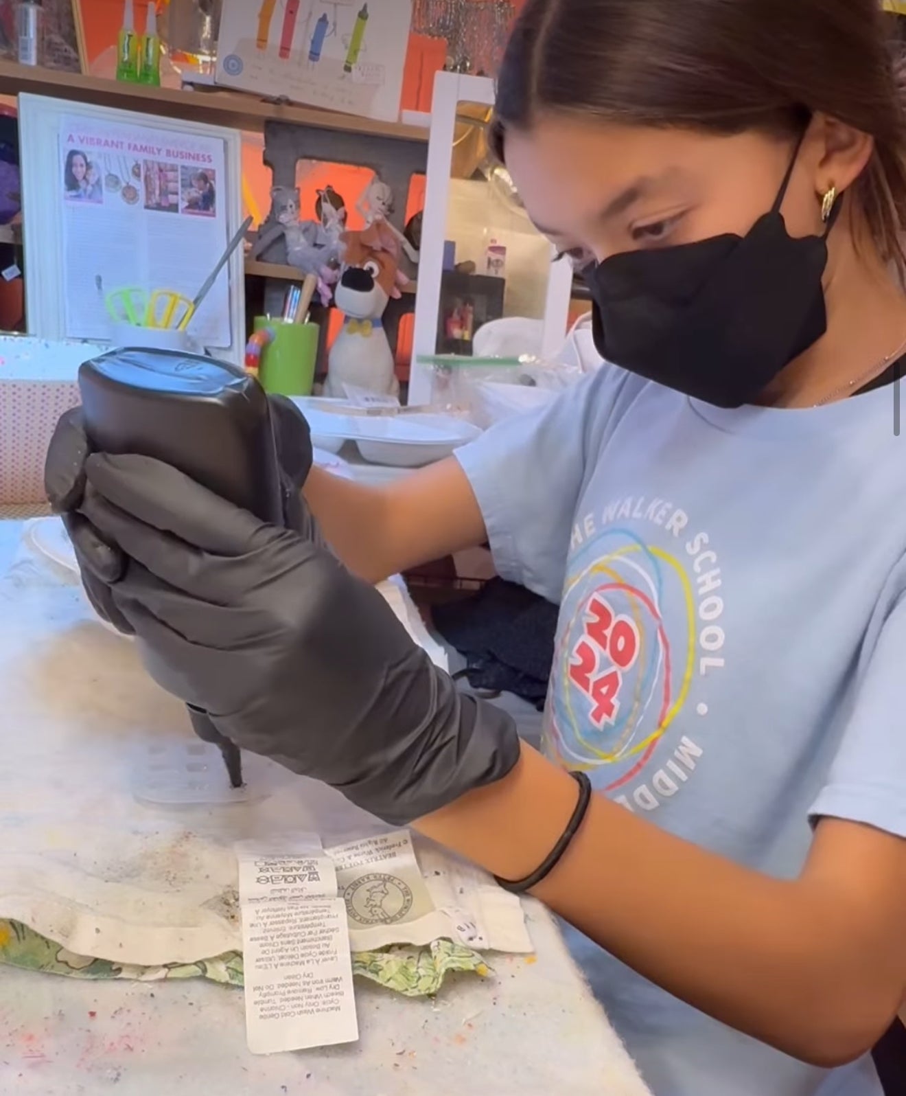 Person wearing a face mask and gloves holding a device in a classroom setting.