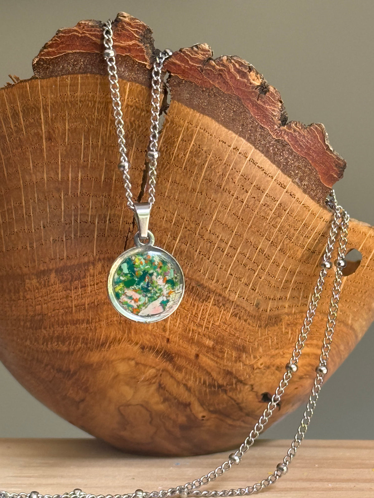 Dime-sized round pendant necklace with ocean blue green orange and pink resin art hanging over wooden bowl, bright gift for her. Crayon jewelry
